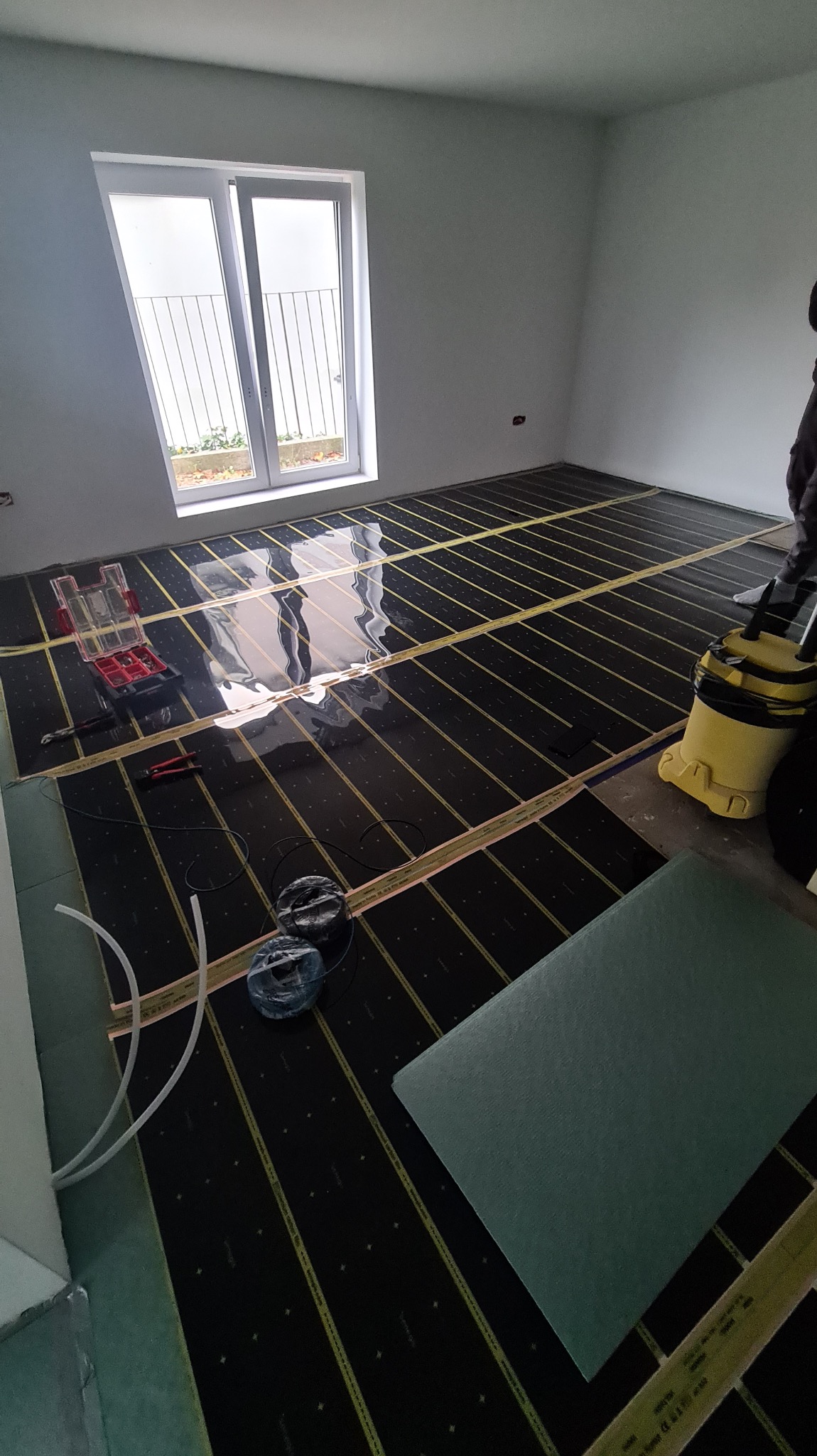 Montaż ogrzewania podłogowego: czarne maty grzewcze z żółtymi paskami ułożone w pokoju z oknem. Widoczne narzędzia, rurki i materiały izolacyjne.