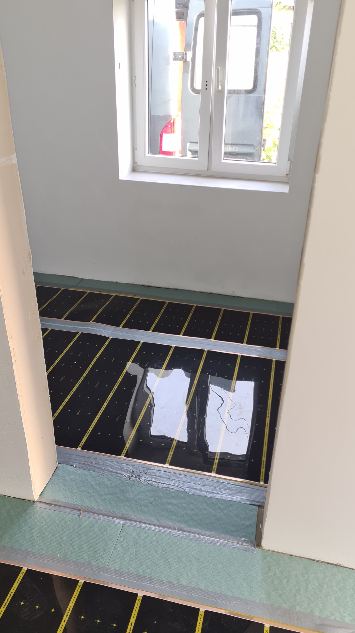 Montaż folii grzewczej pod podłogą w pokoju z oknem. Widoczne czarne panele z żółtymi paskami, białe ściany i szary samochód za oknem.