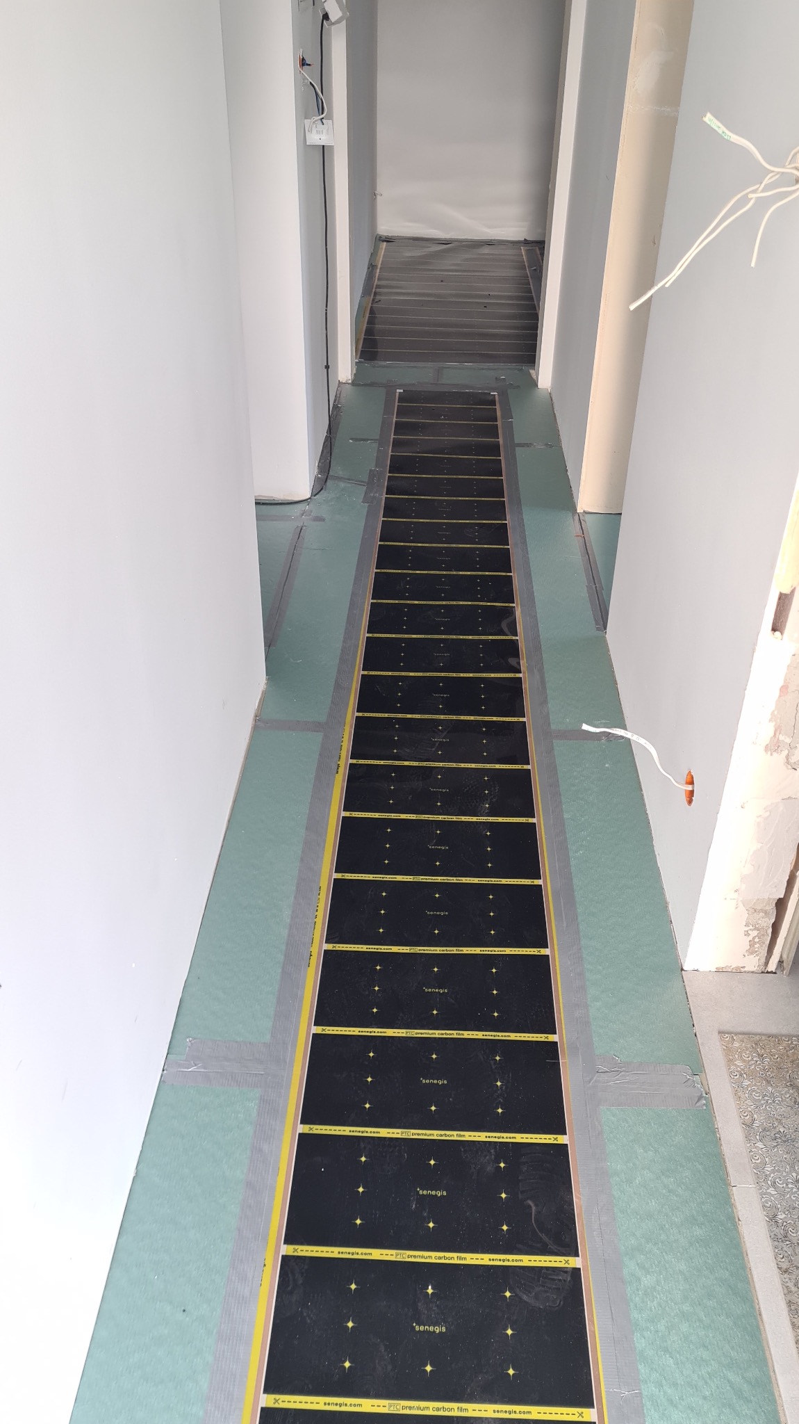 Montaż folii grzewczej Senegis na podłodze w korytarzu, widoczne kable i taśmy montażowe. Prace wykończeniowe w trakcie realizacji.