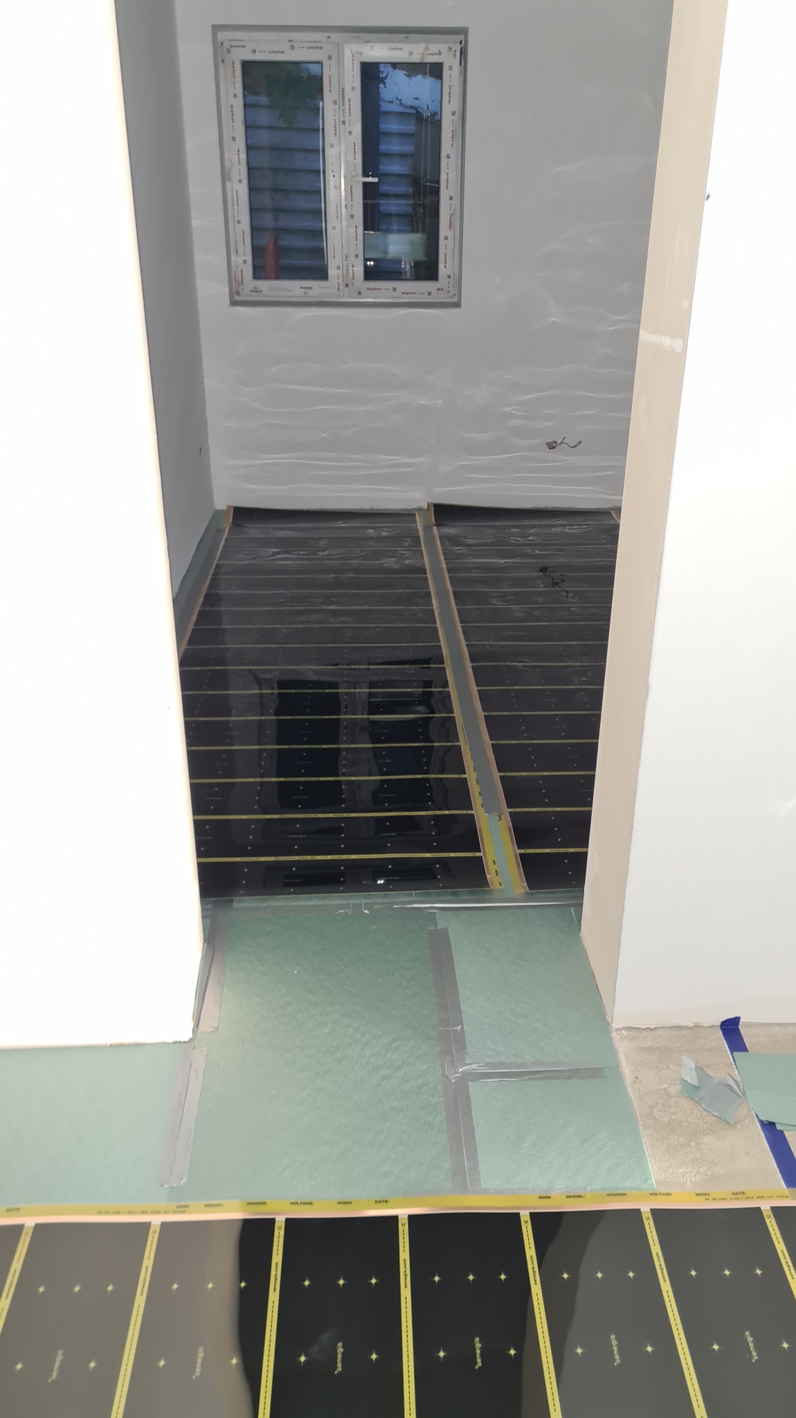 Montaż folii grzewczej na podłodze wewnątrz pomieszczenia z oknem. Widoczne elementy instalacji i warstwy izolacyjne. Etap prac przed wykończeniem podłogi.