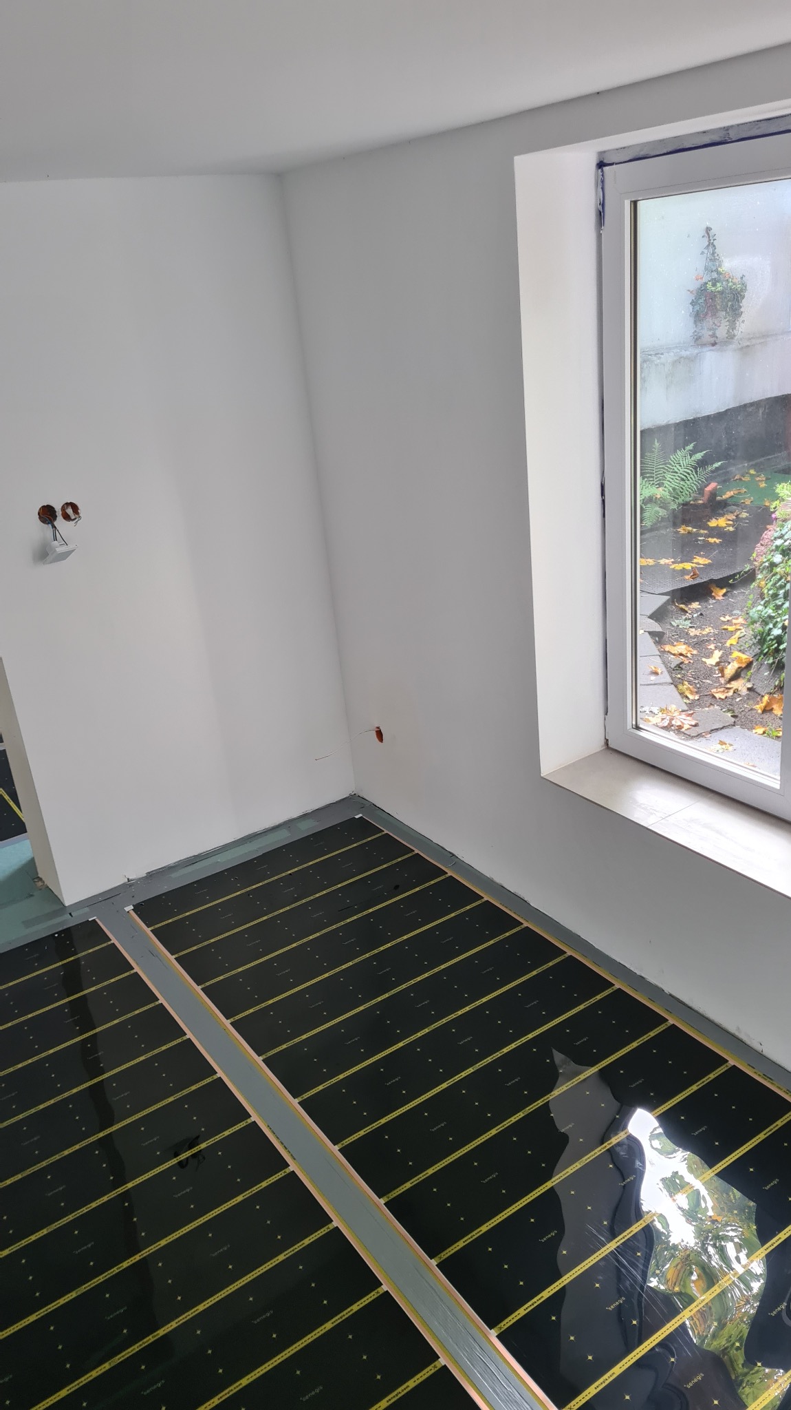 Montaż folii grzewczej na podłodze w jasnym pomieszczeniu z oknem. Widoczne kable elektryczne wystające ze ściany. Układ folii równoległy do okna.