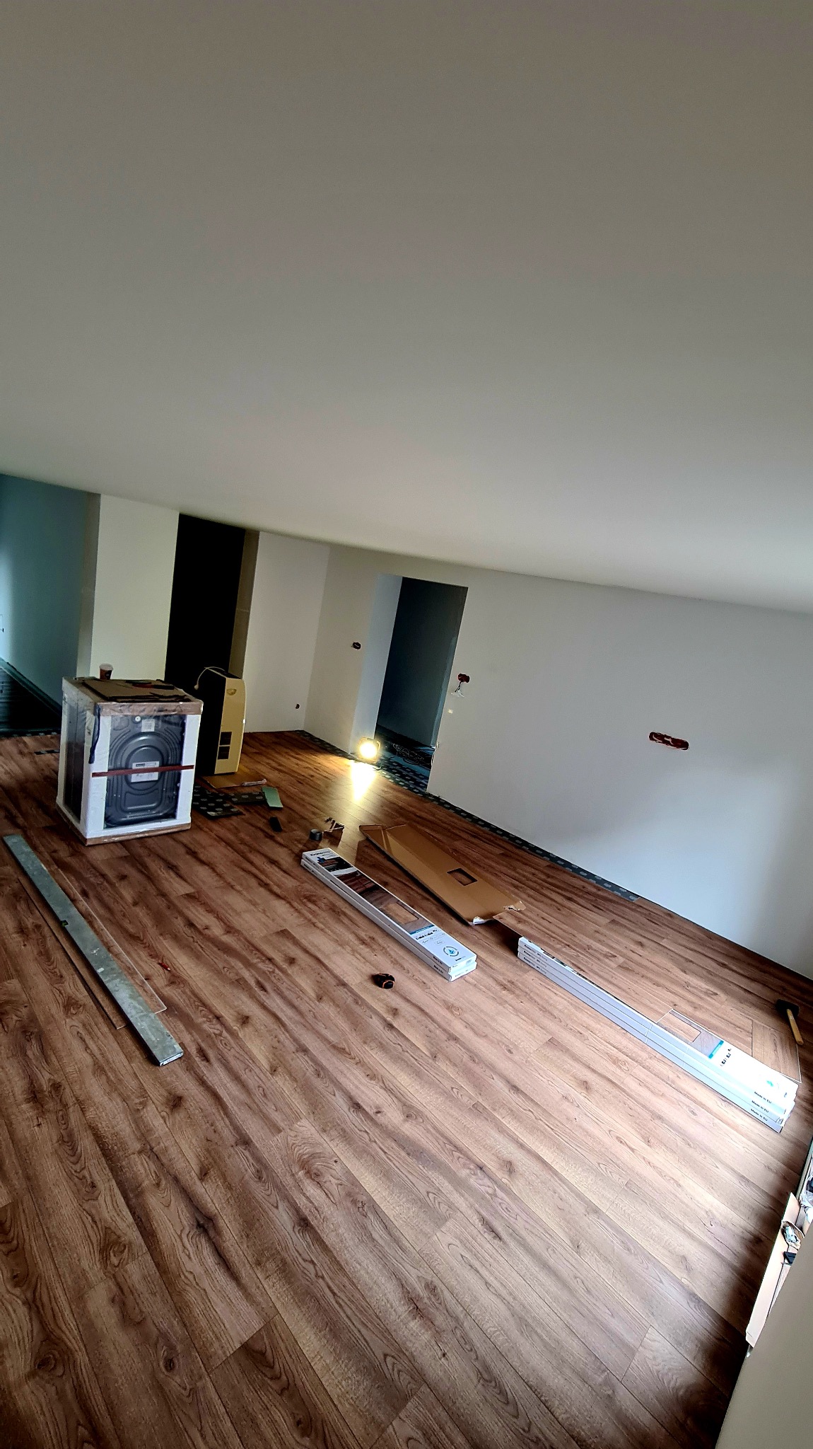 Montaż paneli podłogowych w jasnym pokoju z białymi ścianami. Widoczne narzędzia, opakowania paneli i fragment grzejnika. Podłoga w odcieniach brązu imitująca drewno.