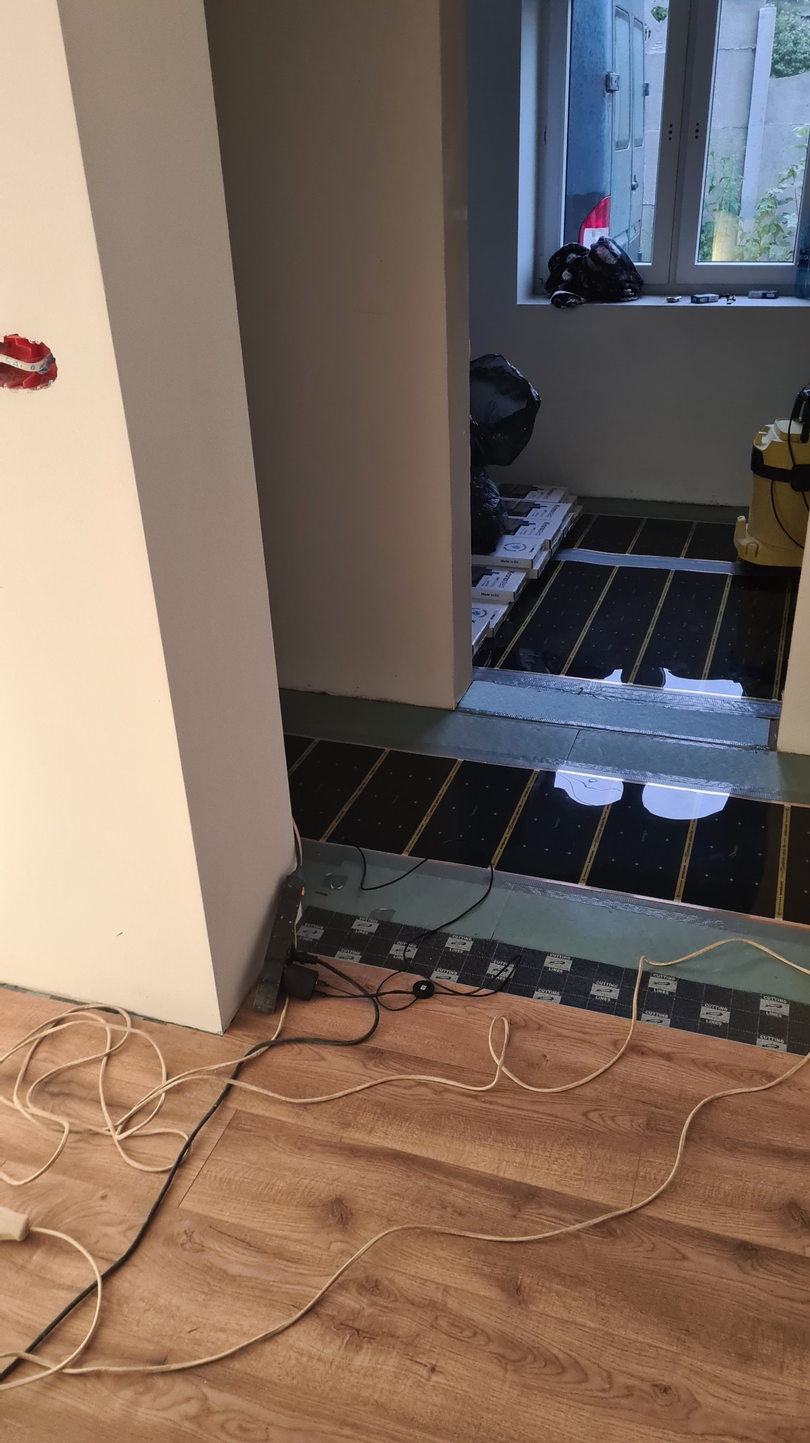 Montaż ogrzewania podłogowego: czarne maty grzewcze ułożone na podłożu, widoczne kable i częściowo ułożona podłoga z paneli drewnopodobnych. Prace w toku.