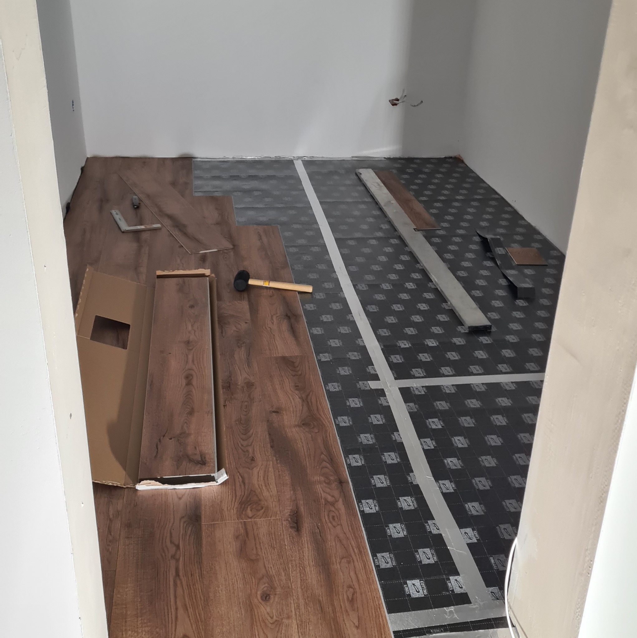 Układanie paneli podłogowych w pokoju. Widoczne narzędzia: młotek, kątownik, listwa. Część podłogi pokryta podkładem, część panelami o wzorze drewna.