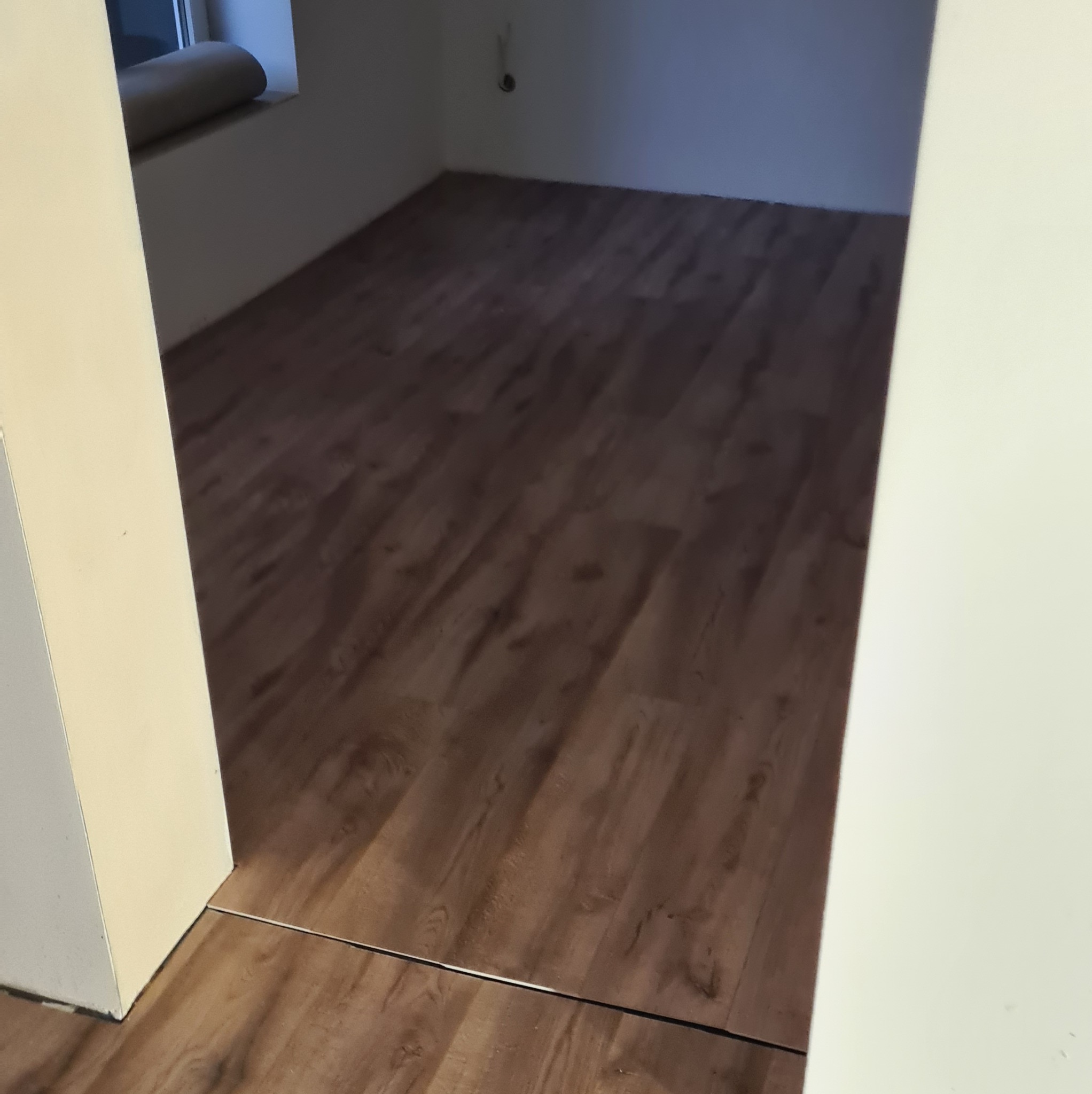 Układanie paneli podłogowych w pokoju, widok zza progu. Nowa, drewniana podłoga w ciepłym odcieniu brązu. Widoczne łączenia paneli i listwa progowa.