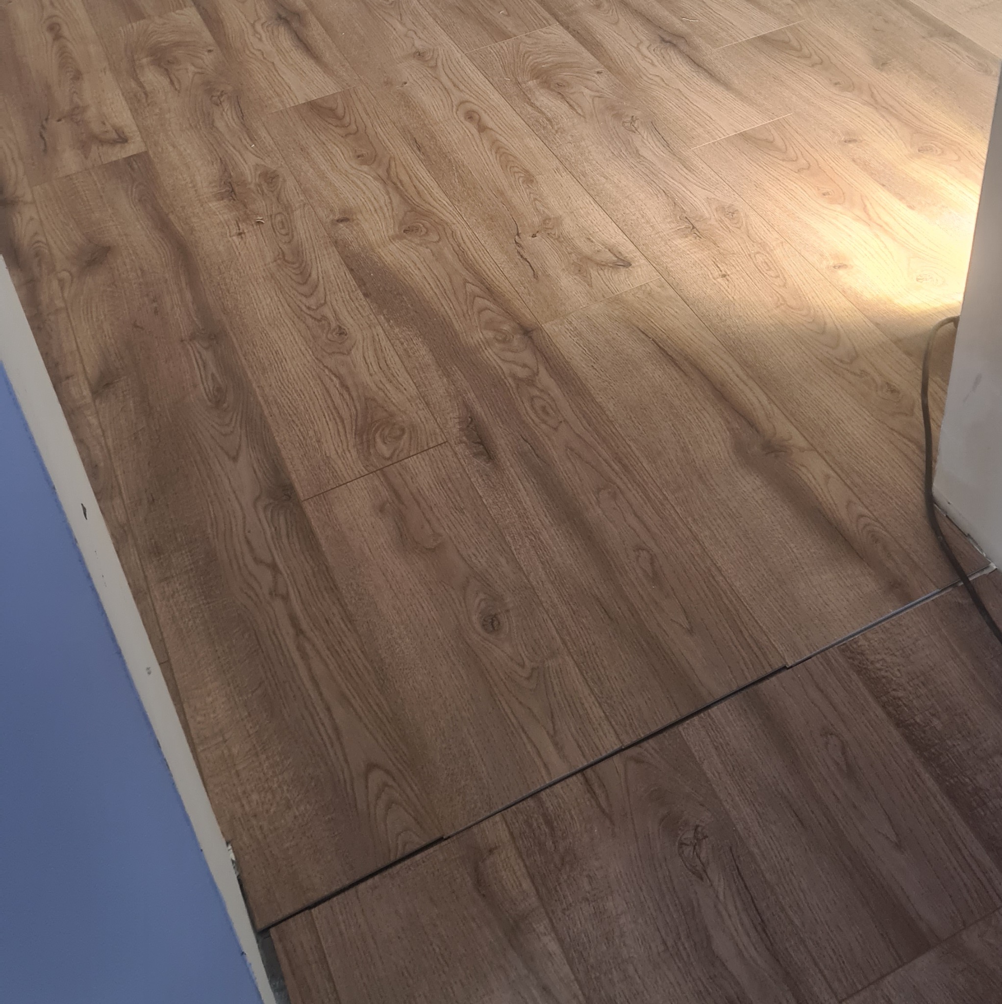 Ukończona podłoga z paneli drewnopodobnych, ułożonych diagonalnie. Widoczne łączenia między panelami i fragment niebieskiej ściany przy krawędzi. Padające światło podkreśla strukturę drewna.