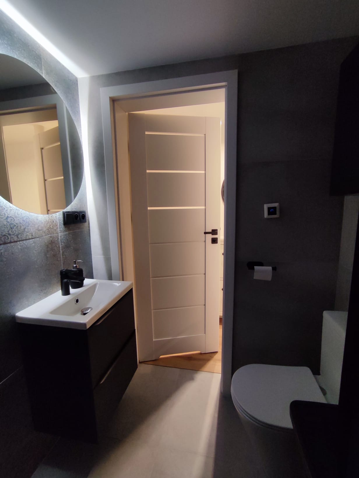 Nowoczesna łazienka z okrągłym lustrem podświetlanym LED, czarną szafką pod umywalką i białą toaletą. Minimalistyczny design z szarymi płytkami i białymi drzwiami.