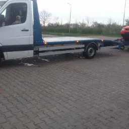 Biała laweta z czerwonym Audi na przyczepie, zaparkowana na brukowanej powierzchni; pies w kabinie kierowcy.