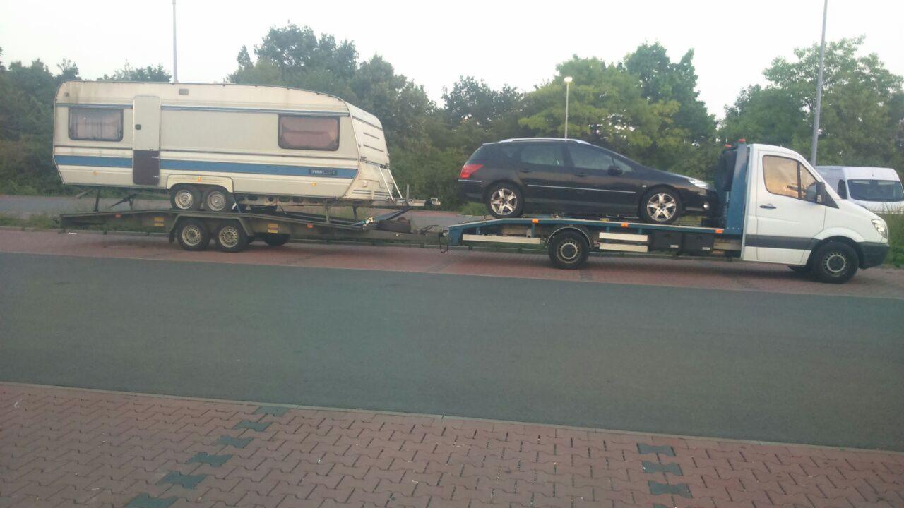 Laweta przewozi jednocześnie samochód osobowy i przyczepę kempingową na dwóch oddzielnych platformach, widok z boku na szarym tle drogi i drzew.