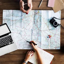 Widok z góry na drewniany stół z rozłożoną mapą, laptopem z otwartą wyszukiwarką, notatnikiem, aparatem fotograficznym, filiżanką kawy i zaznaczonymi pinezkami, sugerujący planowanie podróży...