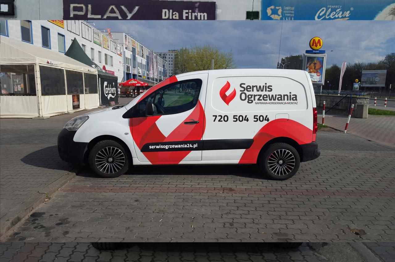 Biały samochód dostawczy z czerwonymi elementami graficznymi, logo firmy 'Serwis Ogrzewania' i numerem telefonu, zaparkowany na brukowanym placu miejskim z budynkami w tle.