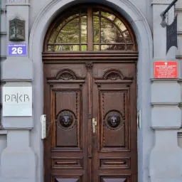 Wejście do kancelarii prawnej z masywnymi, drewnianymi drzwiami ozdobionymi płaskorzeźbami lwów, adres '26' i tabliczka z nazwą kancelarii po lewej stronie, tablica informacyjna komornika sądowego...