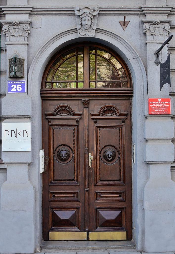 Wejście do kancelarii prawnej z masywnymi, drewnianymi drzwiami ozdobionymi płaskorzeźbami lwów, adres '26' i tabliczka z nazwą kancelarii po lewej stronie, tablica informacyjna komornika sądowego...