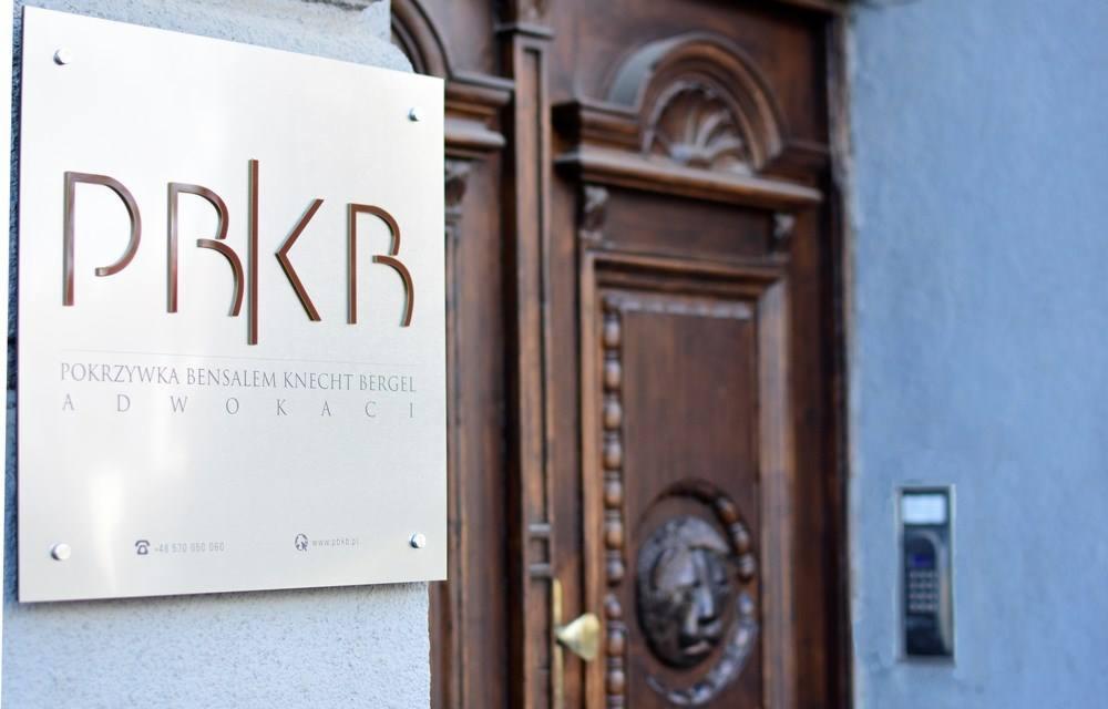 Tabliczka z nazwą kancelarii 'PRKB Pokrzywka Bensalem Knecht Bergel Adwokaci' na srebrnym tle, przymocowana do ściany obok ciemnych, drewnianych drzwi z dekoracyjnym lwem.