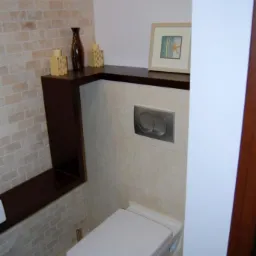 Nowoczesna toaleta podwieszana z chromowanymi przyciskami spłukiwania, umieszczona na tle beżowych płytek imitujących cegłę i jasnych płytek ceramicznych. Na półce nad toaletą dekoracje: ramka ze...