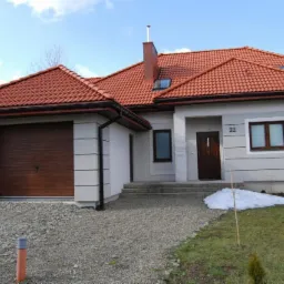 Parterowy dom z garażem, pokryty czerwoną dachówką, z numerem 22 nad drzwiami wejściowymi, na podjeździe żwirowym i trawnikiem z resztkami śniegu.