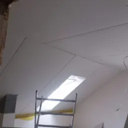 Montaż płyt na poddaszu z oknem dachowym, widoczna drabina i surowe wykończenie ścian.