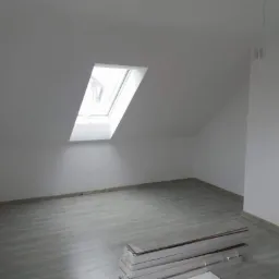Poddasze po remoncie z oknem dachowym, świeżo położone panele podłogowe w odcieniach szarości, białe ściany i sufit, przygotowane paczki paneli w rogu pomieszczenia.