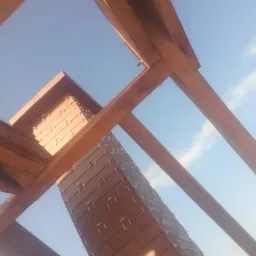 Murowany komin z cegły, widok z dołu przez drewnianą konstrukcję dachu, widoczne krzyżyki dystansowe między cegłami, błękitne niebo w tle.