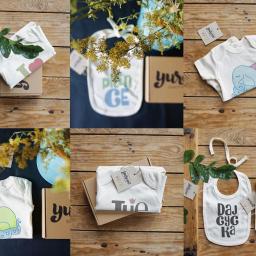 Yurcov - Kompozycja z nadrukami na dziecięcych body i śliniakach marki Yurcov Kids, prezentowanych na tle drewnianych powierzchni i dekoracji roślinnych, w tym gałązek z żółtymi kwiatami i zielonych liści.