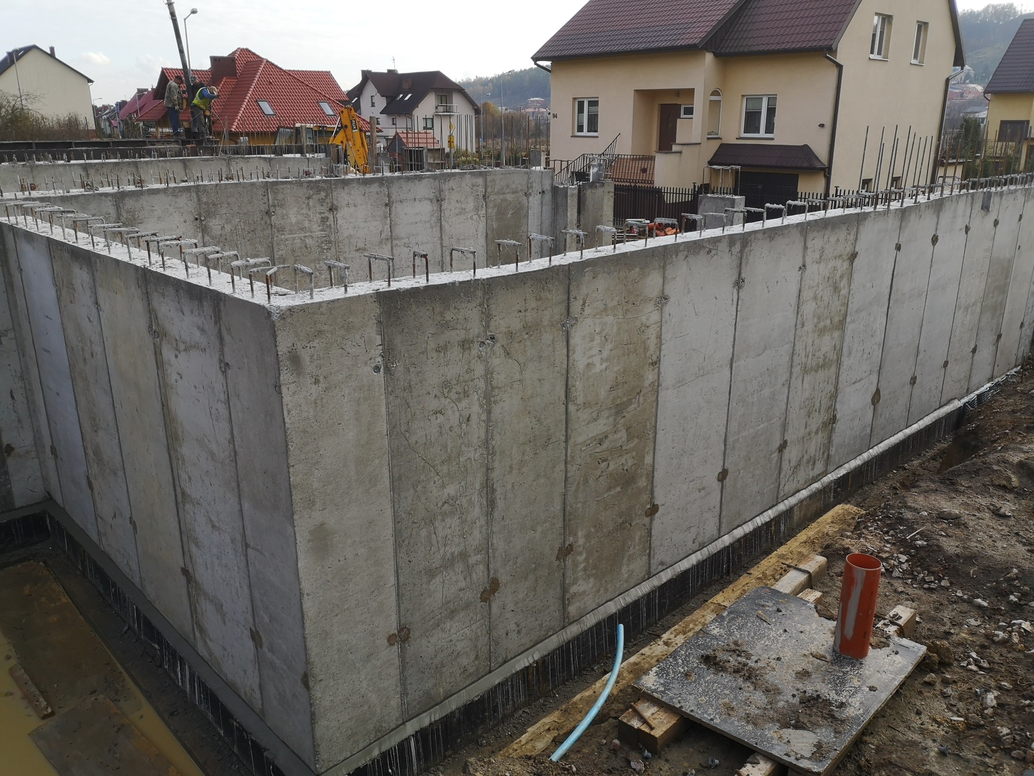 Surowe betonowe fundamenty domu jednorodzinnego z wystającymi prętami zbrojeniowymi, widoczne szalunki, w tle zabudowa mieszkalna.