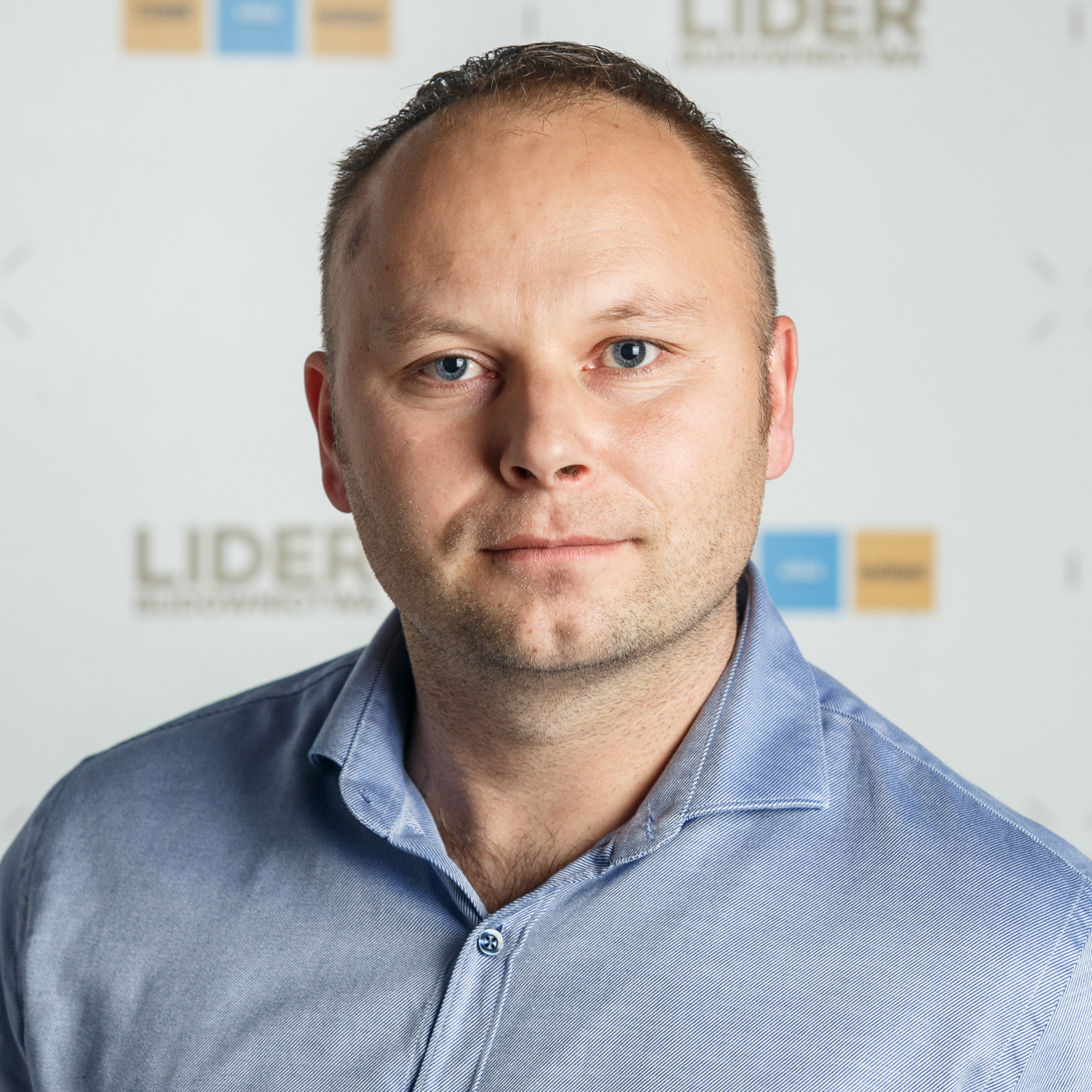 Portret mężczyzny w niebieskiej koszuli na jasnym tle z logo 'Lider Budownictwa'. Umiarkowane oświetlenie, skupienie na twarzy i wyrazie oczu.