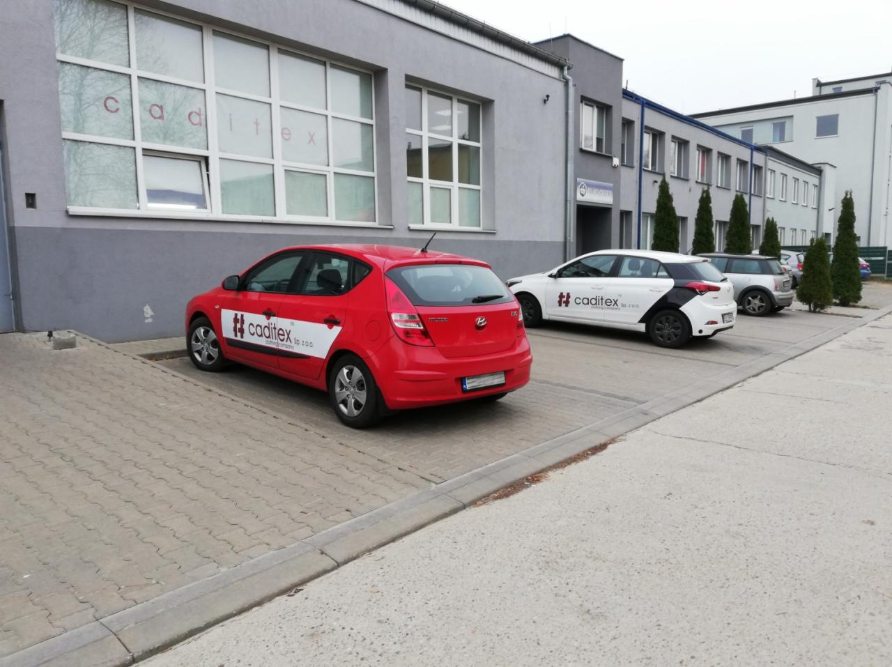 Czerwony samochód z logo Caditex parkuje przed budynkiem firmy odzieżowej z widocznym napisem w oknach, obok zaparkowany biało-czarny samochód z tym samym logo.