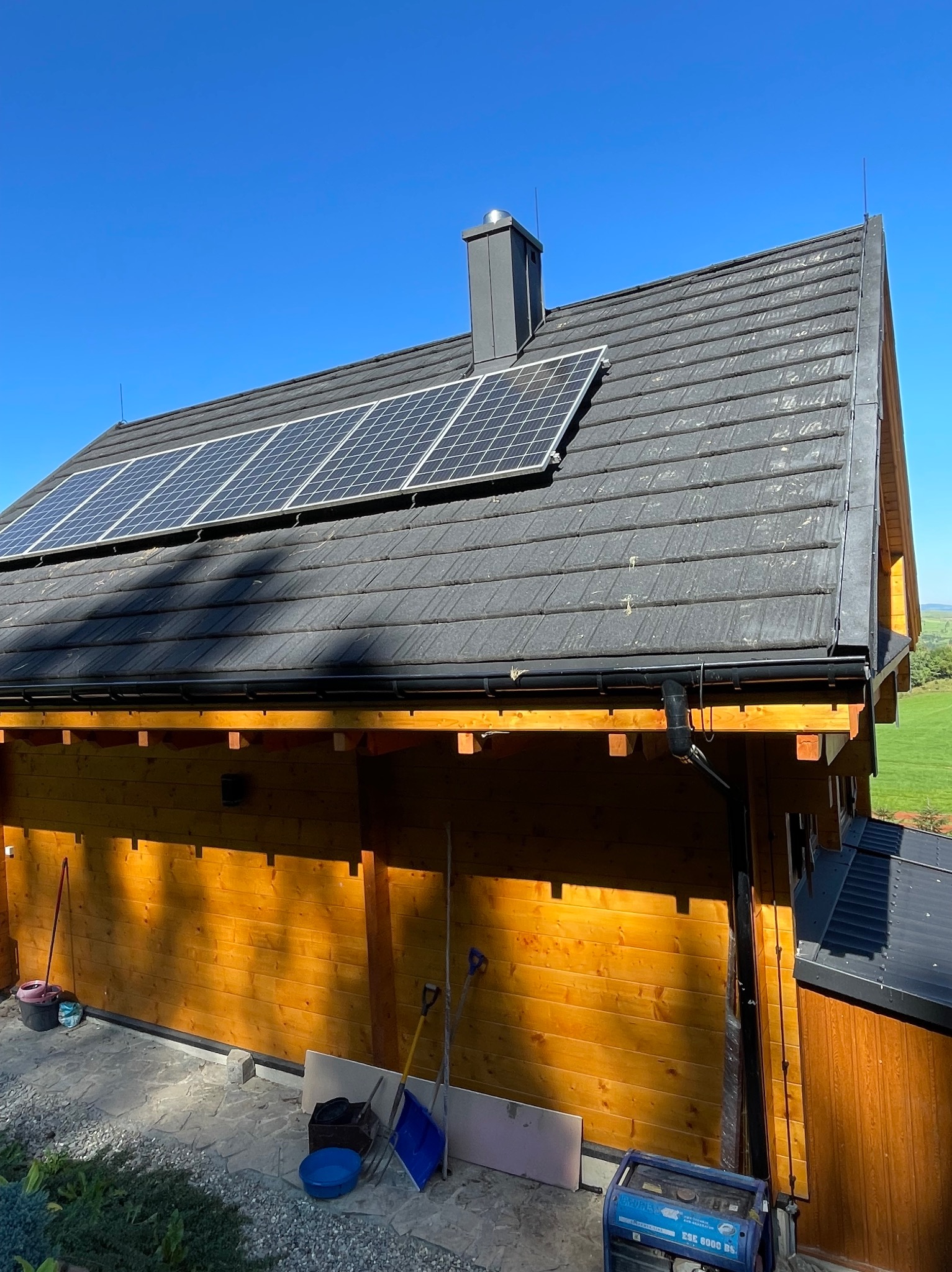 Dom z panelami słonecznymi na dachu z dachówki ceramicznej, widoczne instalacje odgromowe, generator prądu na zewnątrz budynku i narzędzia budowlane.