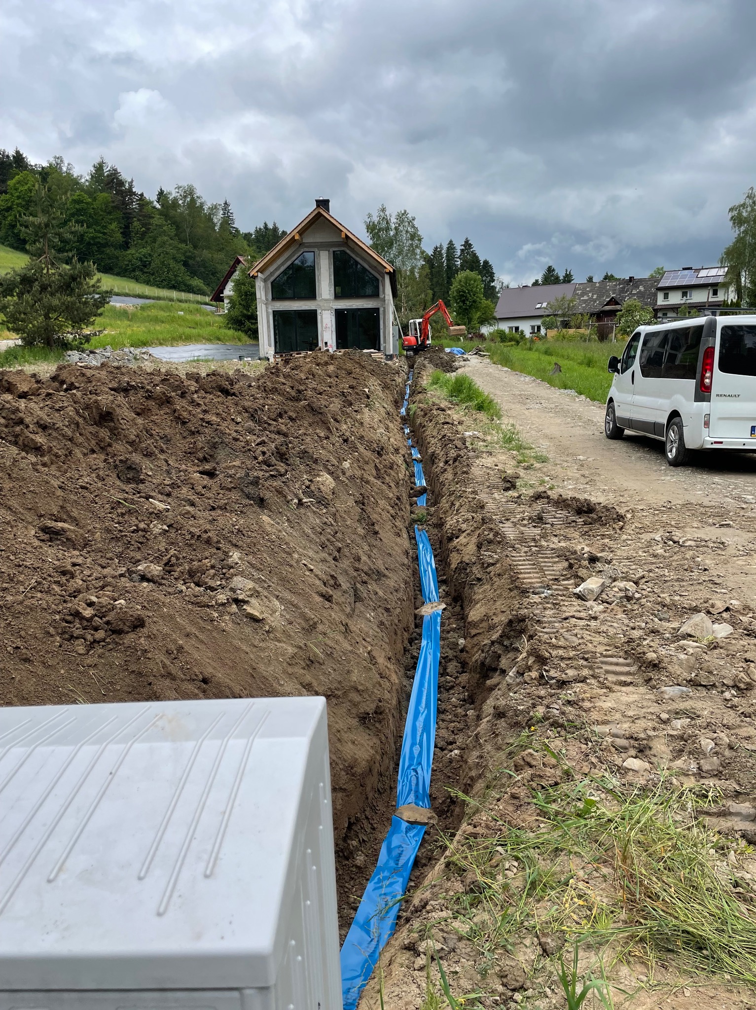 Wykop z niebieską folią zabezpieczającą rury, widoczny biały samochód dostawczy Renault i koparka w tle, dom w stanie surowym otwartym.