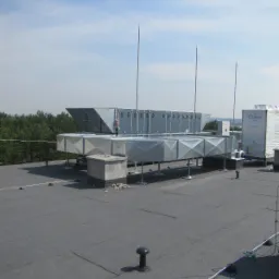 Instalacja odgromowa na dachu budynku, widoczne metalowe maszty i elementy przewodzące, tło niebieskie niebo.