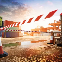 Strefa magazynowa z kontenerami i wózkiem widłowym, oznaczona taśmą ostrzegawczą i znakiem 'Uwaga! Ruch pojazdów', mokra nawierzchnia odbija promienie zachodzącego słońca.