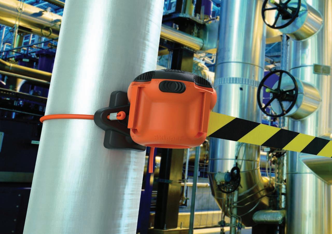 Pomarańczowy moduł barierki ochronnej Skipper XS przymocowany do srebrnej rury w hali przemysłowej, z widoczną czarno-żółtą taśmą ostrzegawczą.