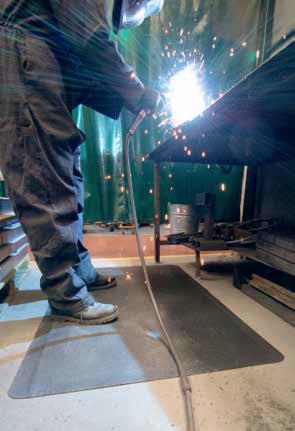 Spawacz w kombinezonie i masce ochronnej spawa metalowy element, iskry, stanowisko pracy z matą antypoślizgową.