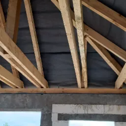 Wnętrze domu w stanie surowym otwartym: widoczne drewniane elementy konstrukcji dachu na tle czarnej membrany dachowej, ściany z betonu komórkowego i nadproża okienne.