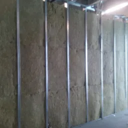 Montaż izolacji z wełny mineralnej w metalowej konstrukcji szkieletowej ściany wewnętrznej, widok z boku na fragment robót w słabym oświetleniu.