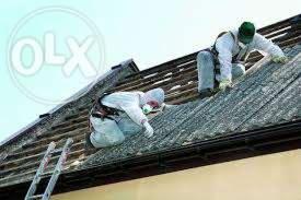 Dwóch pracowników w kombinezonach ochronnych i maskach na dachu podczas pracy, widoczna drabina oparta o ścianę budynku, jasne niebo w tle.