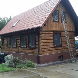 Drewniany dom z czerwoną dachówką, okna zabezpieczone folią malarską, przystawiona aluminiowa drabina, widoczne ślady świeżo nałożonej bejcy na elewacji.