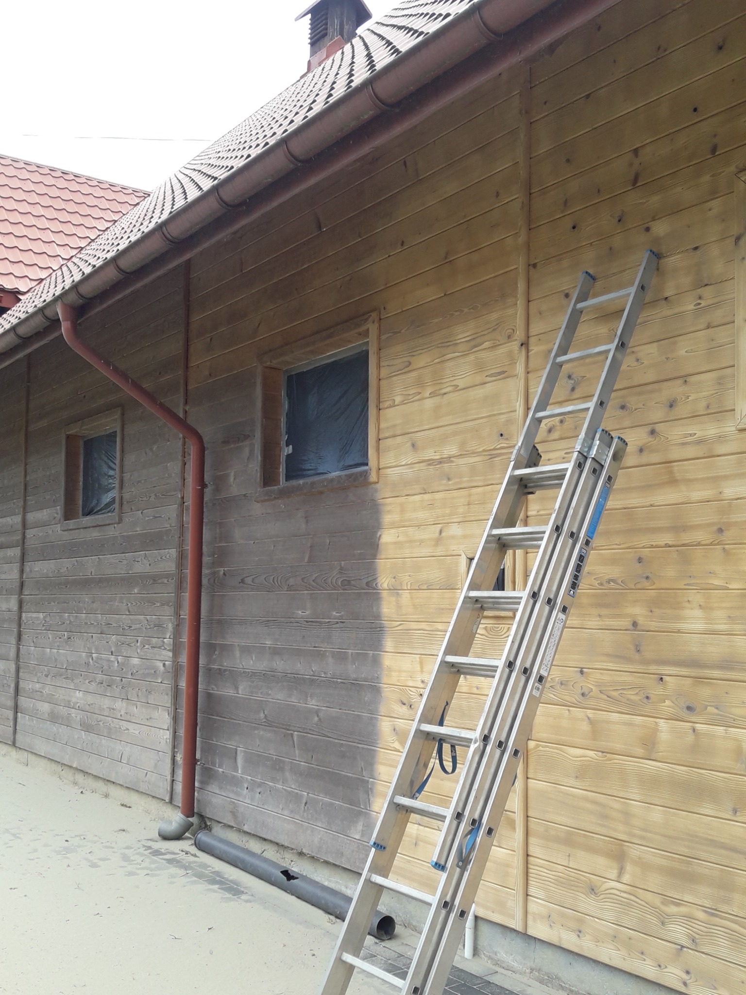 Drewniana elewacja budynku w trakcie renowacji, z widoczną różnicą w kolorze drewna po lewej i prawej stronie drabiny, okna zabezpieczone folią.