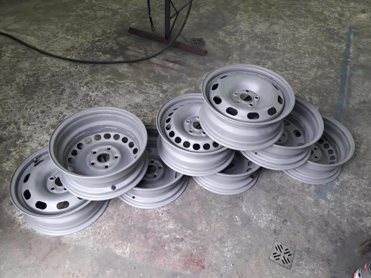 Stos srebrnych felg samochodowych z otworami wentylacyjnymi, ułożonych na betonowej podłodze w warsztacie, po procesie malowania.