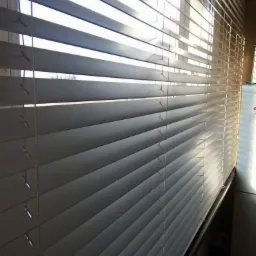 Zamknięte poziome osłony okienne w jasnym kolorze, widoczne sznurki sterujące, zza okna przebija światło słoneczne.