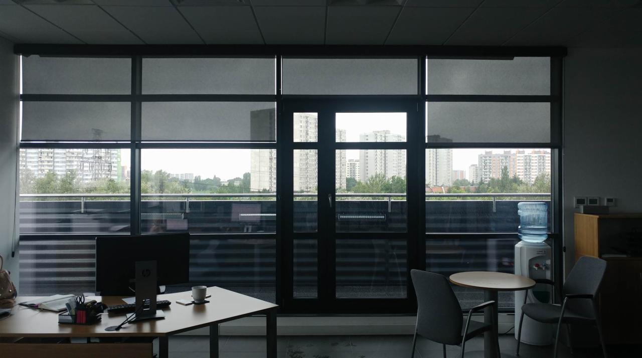 Biuro z dużymi oknami i szarymi, zaciemniającymi roletami zwiniętymi do połowy wysokości, widok na miasto w tle.