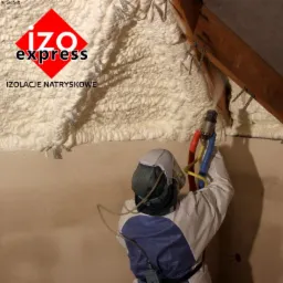 Pracownik w kombinezonie ochronnym aplikuje izolację natryskową na poddaszu, widoczne krokwie i metalowe mocowania.