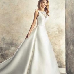 Śliczne Suknie Ślubne - Modelka w białej sukni ślubnej z koronkową górą i satynowym dołem, prezentująca kolekcję 'Vanilla Sposa' na tle teksturowanej ściany.