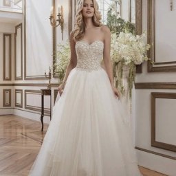 Śliczne Suknie Ślubne - Panna młoda w białej, balowej sukni bez ramiączek z bogato zdobionym gorsetem, pozująca w eleganckim wnętrzu z dekoracyjnymi sztukateriami i bukietami białych kwiatów.