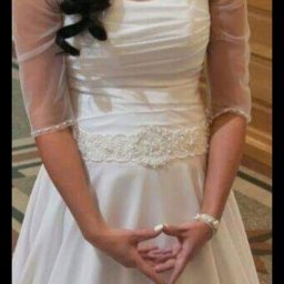 Śliczne Suknie Ślubne - Panna młoda w białej sukni ślubnej z tiulowymi rękawami i ozdobnym pasem, dłonie złożone w charakterystyczny sposób, widoczny srebrny naszyjnik i bransoletka.