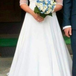 Śliczne Suknie Ślubne - Panna młoda w białej sukni ślubnej z bukietem niebiesko-białych kwiatów, z półprzezroczystymi rękawami, stojąca obok mężczyzny w granatowym garniturze.