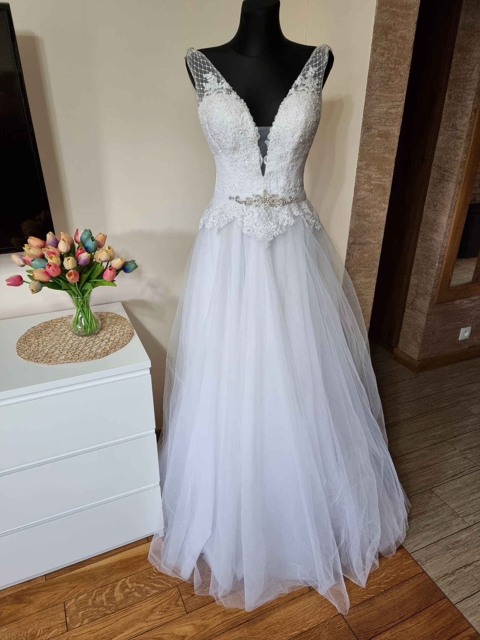 Elegancka suknia ślubna z koronkowym gorsetem i tiulowym dołem, prezentowana na manekinie obok wazonu z kolorowymi tulipanami na białej komodzie.