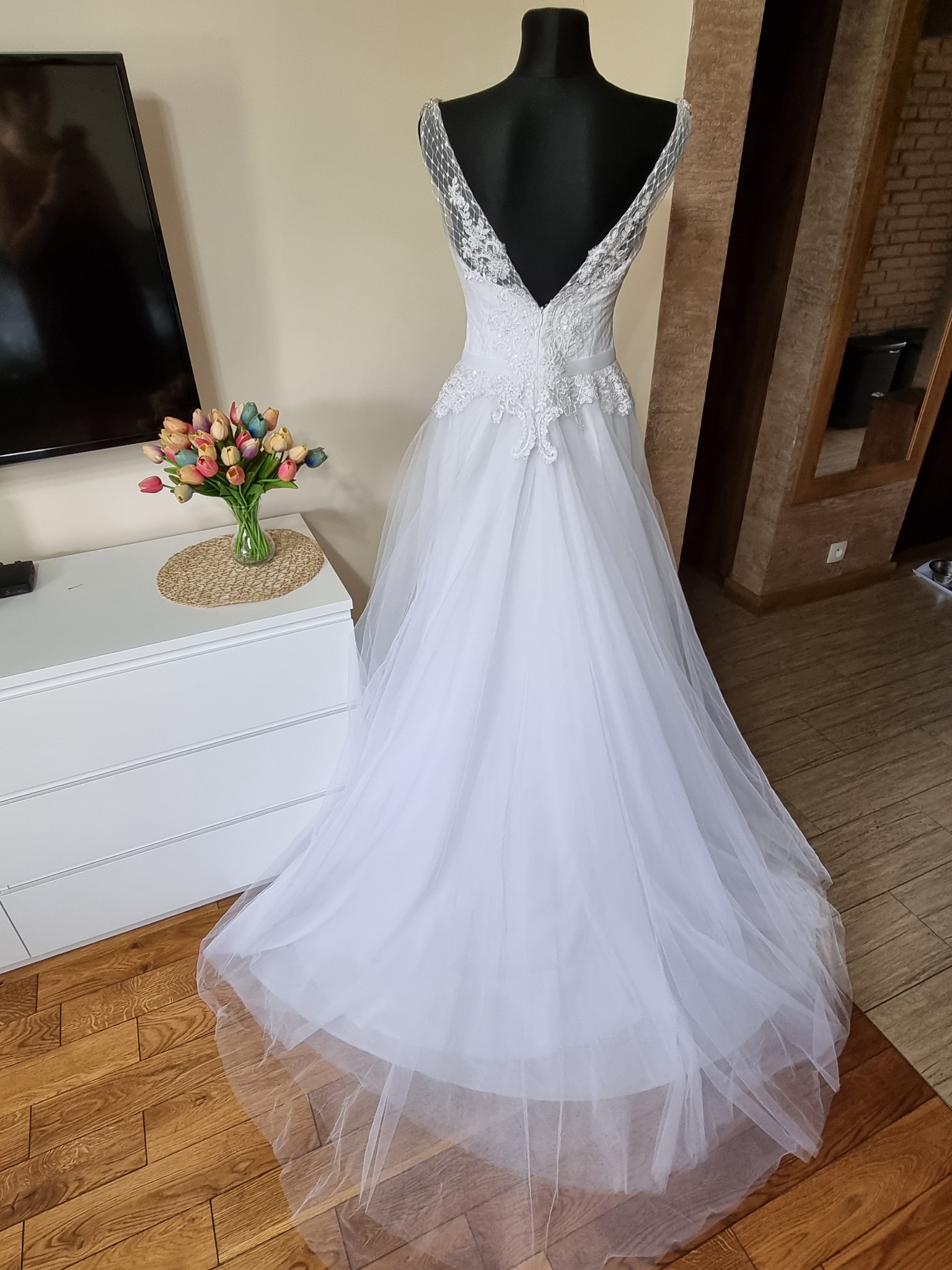 Biała suknia ślubna z koronkowymi zdobieniami na manekinie, z bukietem tulipanów na białej komodzie w tle.