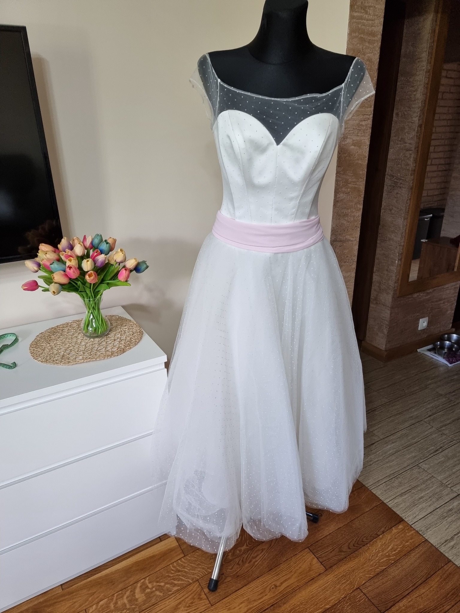 Elegancka suknia ślubna z tiulowym dołem w kropki i różowym pasem, prezentowana na manekinie obok bukietu tulipanów na białej komodzie.