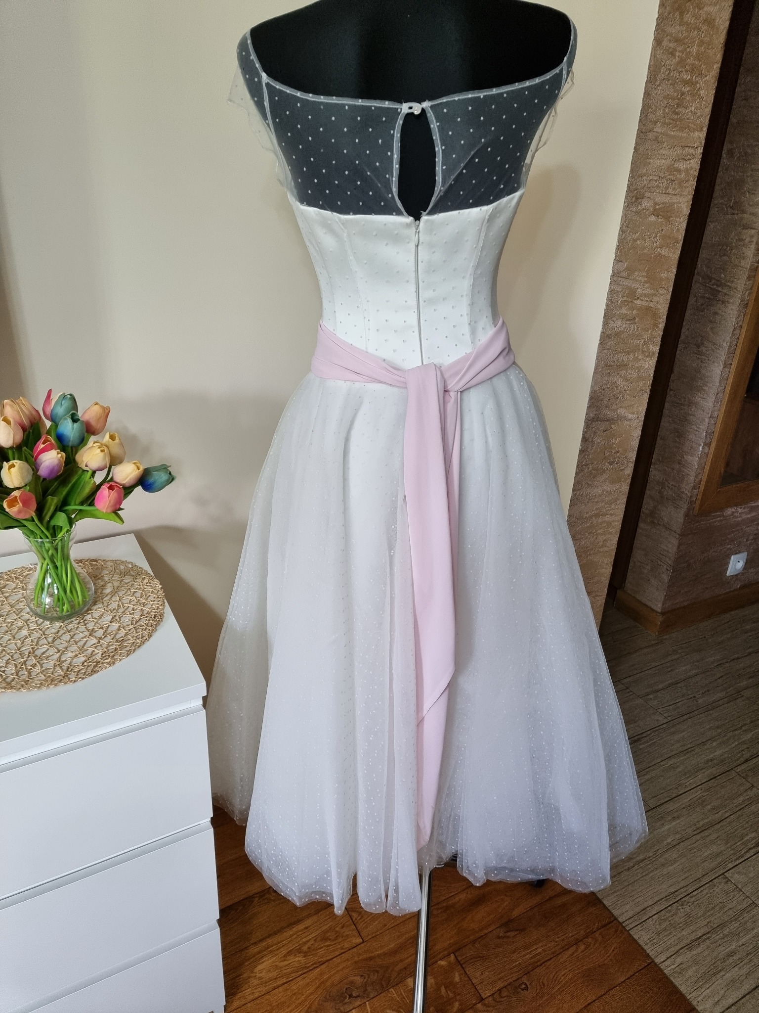 Manekin prezentuje białą sukienkę z tiulową spódnicą w kropki, ozdobioną różową szarfą, obok wazon z kolorowymi tulipanami.